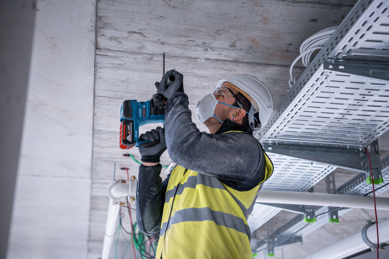 Eine Person mit Sicherheitsausrüstung bohrt auf einer Baustelle in eine Betondecke. Anwendungsbild