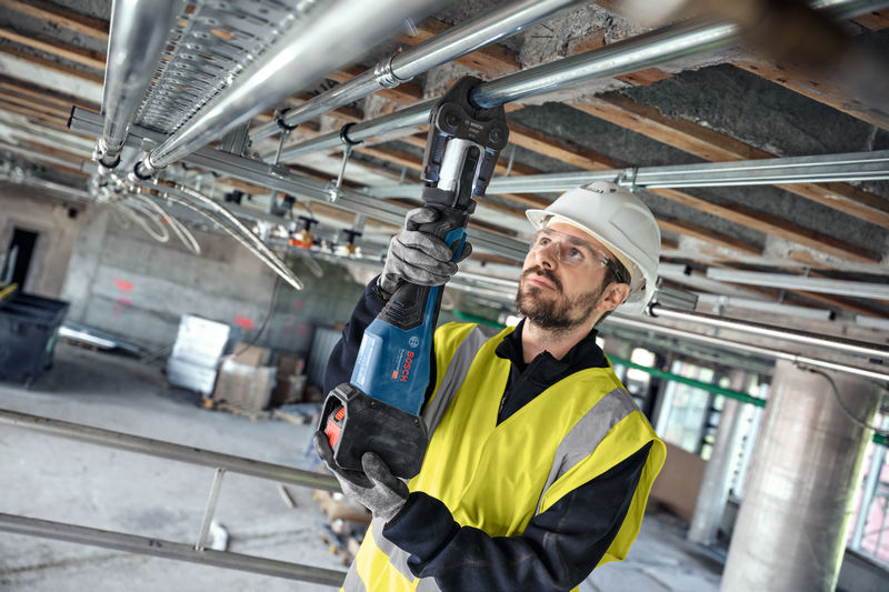 Eine Person mit Sicherheitsausrüstung verwendet ein Akku-Presswerkzeug an Metallrohren über der Decke. Anwendungsbild