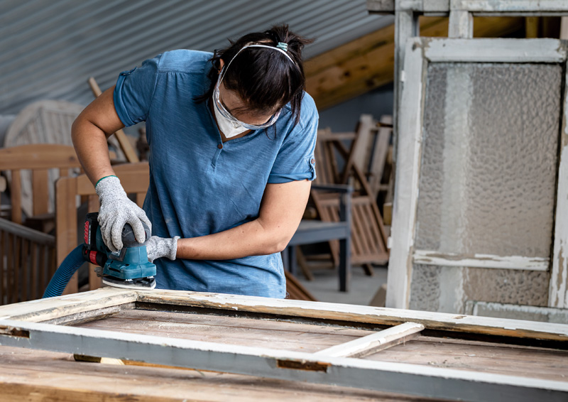 Ein Arbeiter mit Sicherheitsausrüstung schleift einen alten Holzfensterrahmen. Anwendungsbild