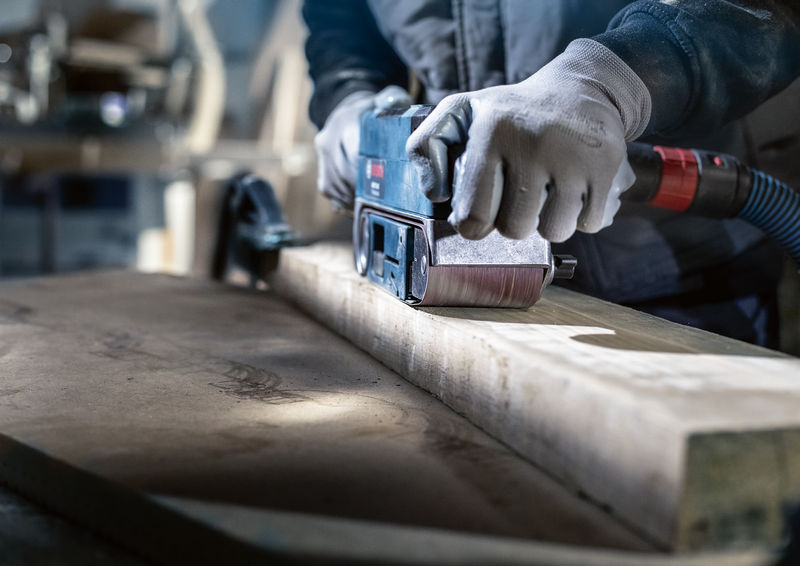 Ein Arbeiter mit Sicherheitsausrüstung schleift einen Holzbalken mit einem Bandschleifer. Anwendungsbild