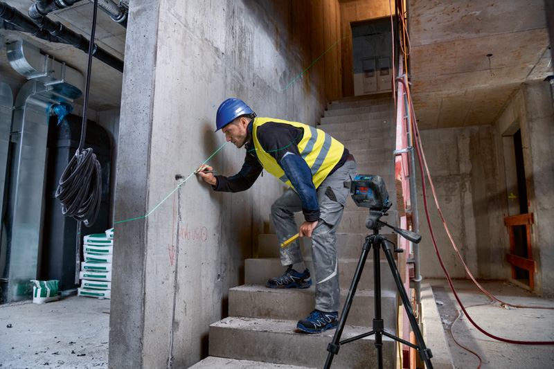 Eine Person mit Sicherheitsausrüstung markiert eine Wand mit einem Lasernivelliergerät auf einem Stativ. Anwendungsbild