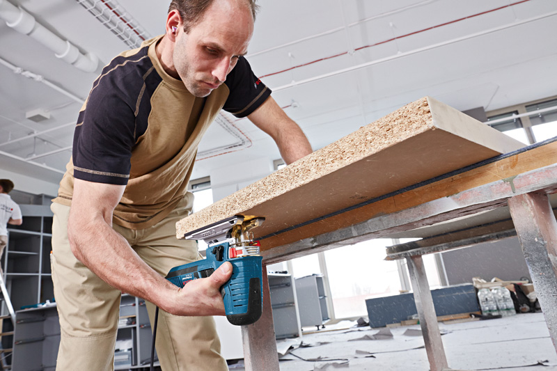 Eine Person mit Schutzausrüstung schneidet mit einer Stichsäge ein Holzbrett auf einer Werkbank zu. Anwendungsbild
