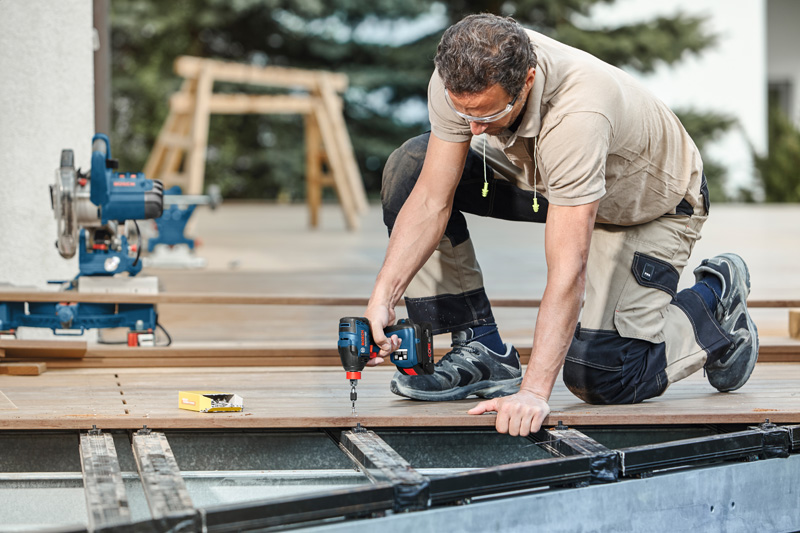 Eine Person mit Sicherheitsausrüstung befestigt Terrassendielen im Außenbereich mit einer Bohrmaschine. Anwendungsbild