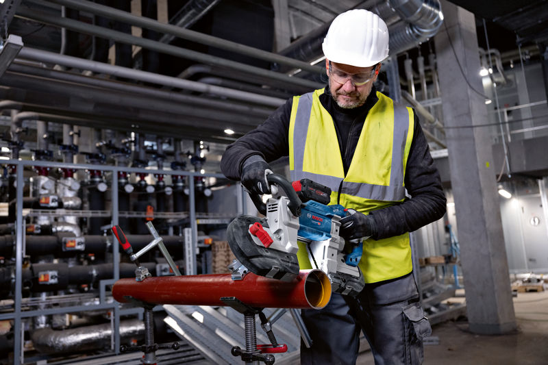 Ein Arbeiter mit Sicherheitsausrüstung schneidet mit einer kabellosen Bandsäge ein Metallrohr. Anwendungsbild