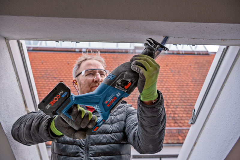 Eine Person mit Schutzausrüstung schneidet mit einer Säbelsäge in einen Fensterrahmen. Anwendungsbild