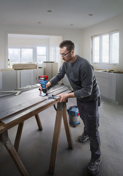 Eine Person mit Sicherheitsausrüstung formt Holz auf einem Arbeitstisch mit einer Oberfräse. Anwendungsbild