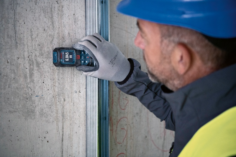 Eine Person mit Sicherheitsausrüstung misst eine Wand mit einem Lasermessgerät. Anwendungsbild