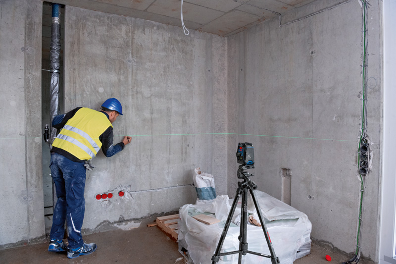 Eine Person mit Sicherheitsausrüstung markiert eine Wand mit einem Lasernivelliergerät auf einem Stativ. Anwendungsbild