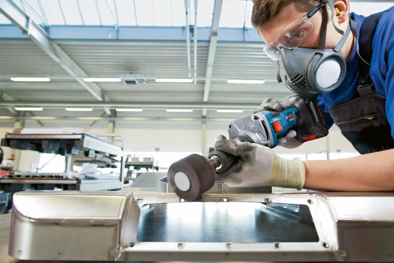 Eine Person mit Schutzausrüstung schleift mit einem Akku-Geradschleifer eine Metalloberfläche. Anwendungsbild