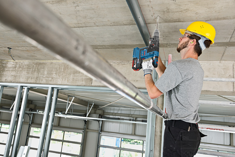 Eine Person mit Sicherheitsausrüstung bohrt auf einer Baustelle in eine Decke. Anwendungsbild