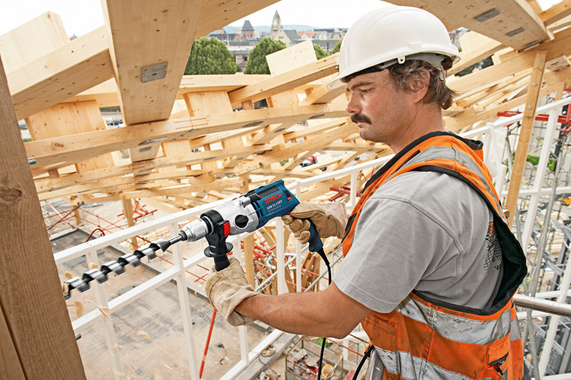 Eine Person mit Sicherheitsausrüstung bohrt auf einer Baustelle in einen Holzbalken. Anwendungsbild