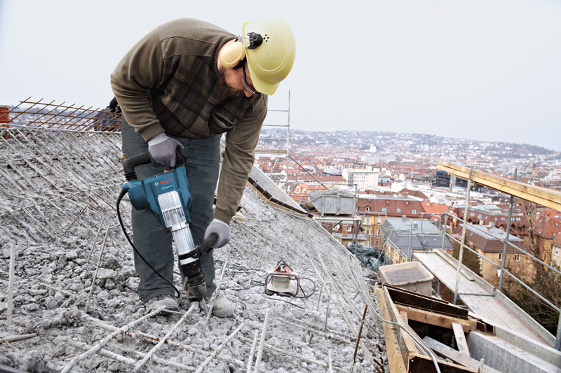 Eine Person mit Sicherheitsausrüstung benutzt einen Abbruchhammer, um Beton auf einem Dach aufzubrechen. Anwendungsbild