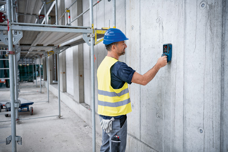 Eine Person mit Sicherheitsausrüstung scannt eine Betonwand mit einem Detektorwerkzeug. Anwendungsbild