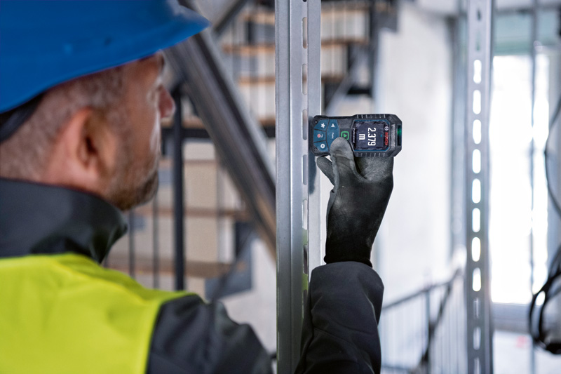 Eine Person mit Sicherheitsausrüstung misst einen Metallträger mit einem Lasermessgerät. Anwendungsbild