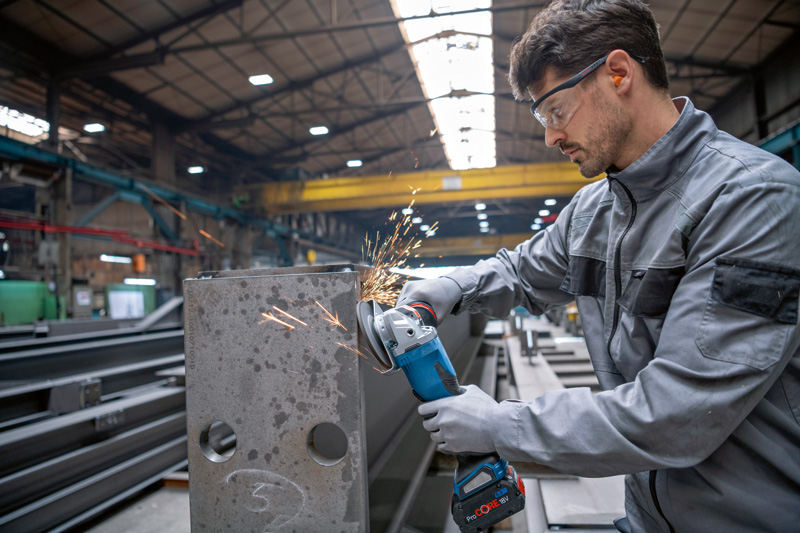Eine Person mit Schutzausrüstung schleift mit einem Akku-Winkelschleifer einen Metallträger. Anwendungsbild