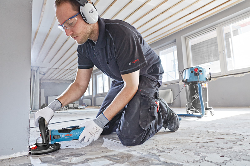 Eine Person mit Sicherheitsausrüstung schleift einen Betonboden mit einem blauen Betonschleifer. Anwendungsbild