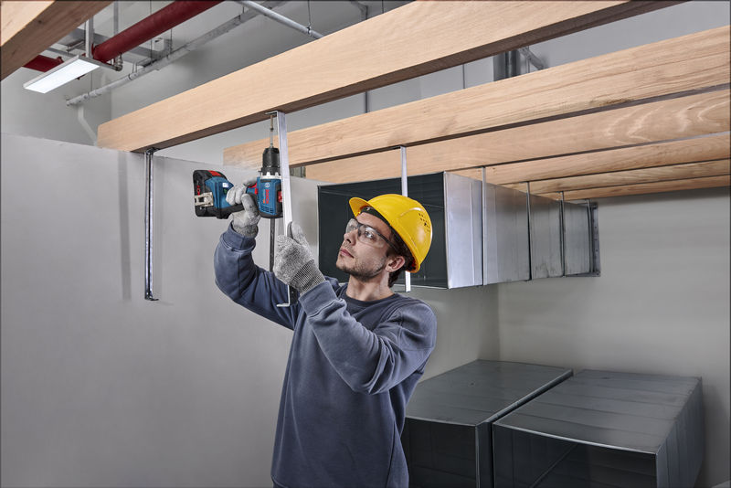 Eine Person mit Sicherheitsausrüstung befestigt mit einer Bohrmaschine Metallstützen über Kopf. Anwendungsbild