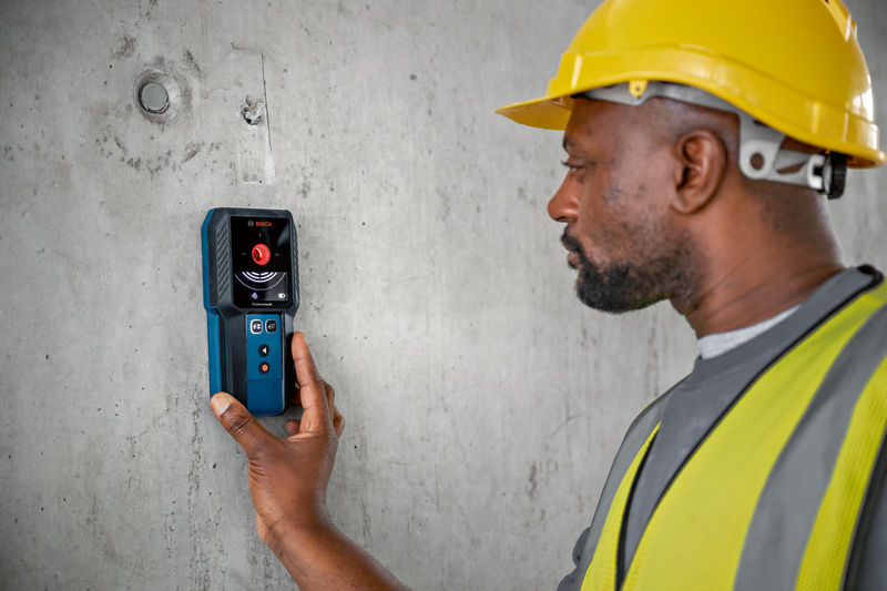 Eine Person mit Sicherheitsausrüstung scannt mit einem Detektor eine Betonwand. Anwendungsbild