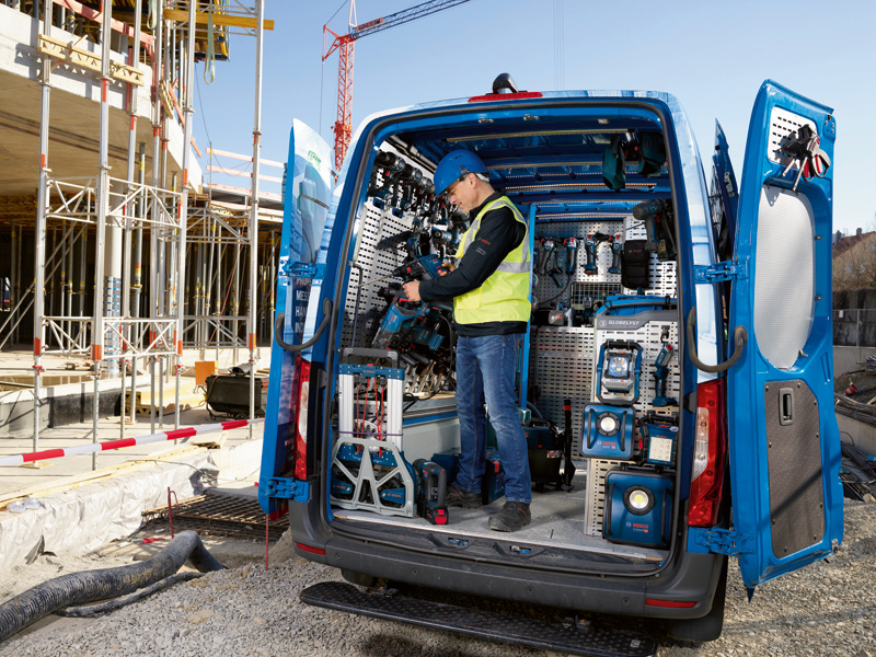 Eine Person in Sicherheitsausrüstung räumt auf einer Baustelle Elektrowerkzeuge in einem Lieferwagen ein. Anwendungsbild