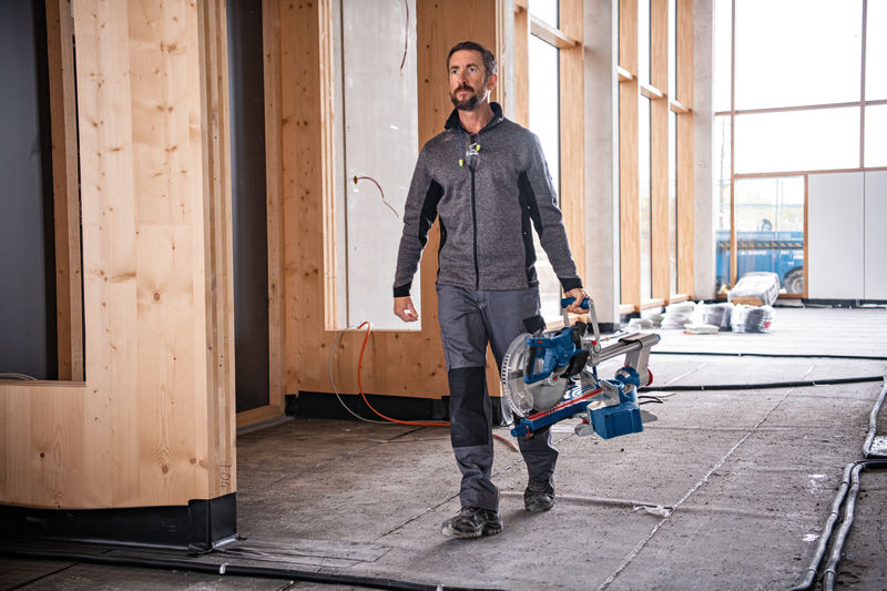 Eine Person mit Sicherheitsausrüstung trägt eine kabellose Gehrungssäge auf einer Baustelle. Anwendungsbild