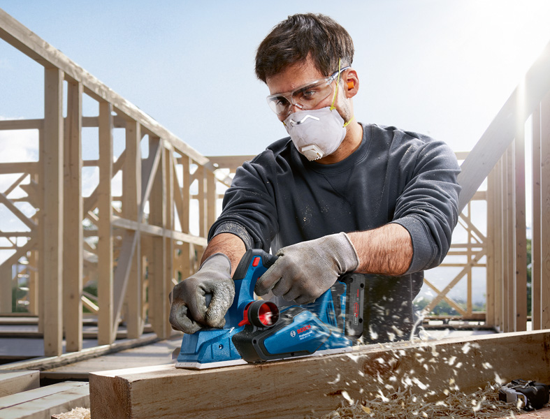 Eine Person mit Sicherheitsausrüstung hobelt auf einer Baustelle einen Holzbalken. Anwendungsbild