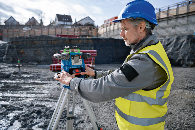Eine Person mit Sicherheitsausrüstung justiert auf einer Baustelle ein Lasernivelliergerät auf einem Stativ. Anwendungsbild