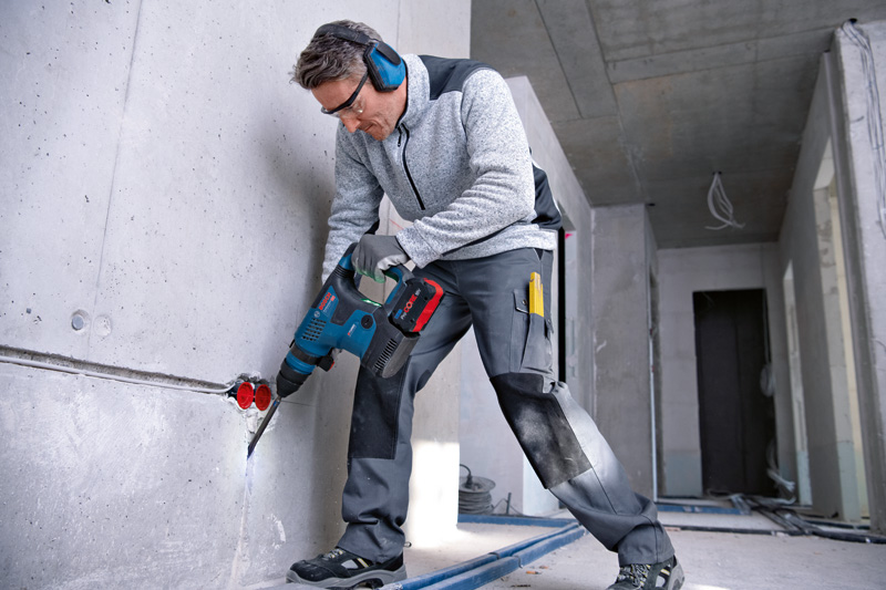 Eine Person mit Sicherheitsausrüstung bohrt in eine Betonwand auf einer Baustelle. Anwendungsbild