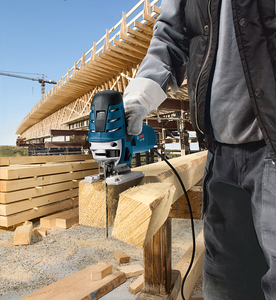 Eine Person mit Sicherheitsausrüstung schneidet auf einer Baustelle mit einer Stichsäge einen Holzbalken durch. Anwendungsbild