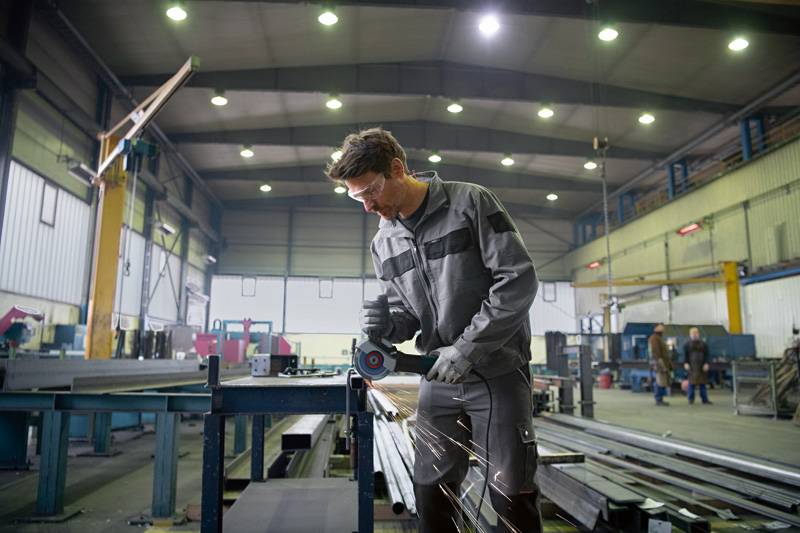 Eine Person mit Schutzausrüstung schleift Metall, Funken fliegen in einer großen Industriewerkstatt. Anwendungsbild
