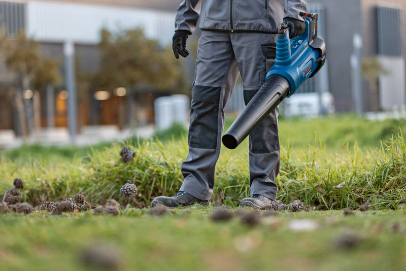 Eine Person mit Sicherheitsausrüstung verwendet einen kabellosen Laubbläser auf Grasboden. Anwendungsbild