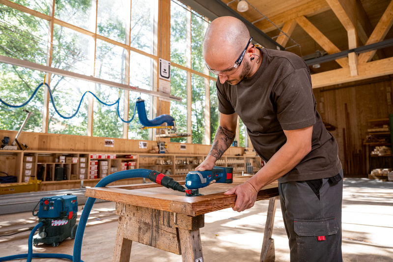 Eine Person mit Schutzausrüstung schleift mit einem Elektrowerkzeug Holz auf einer Werkbank. Anwendungsbild