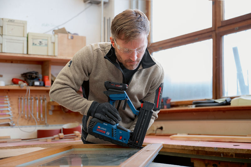 Eine Person mit Sicherheitsausrüstung verwendet einen Akku-Holznagler an einem Fensterrahmen. Anwendungsbild