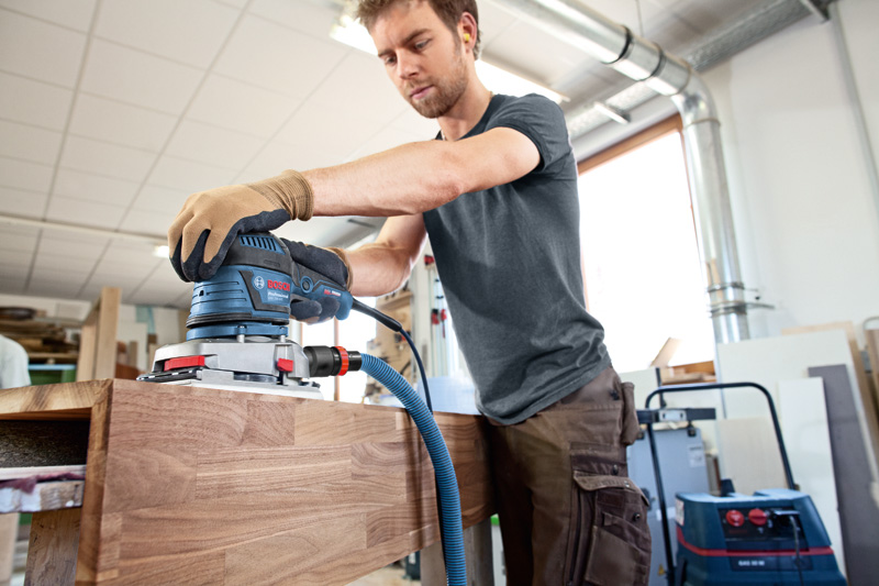 Person mit Schutzausrüstung schleift Holzmöbel mit einem Schwingschleifer. Anwendungsbild