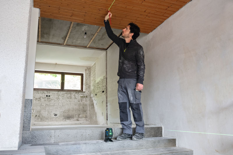 Eine Person mit Sicherheitsausrüstung misst eine Holzdecke mit einem Lasernivelliergerät. Anwendungsbild