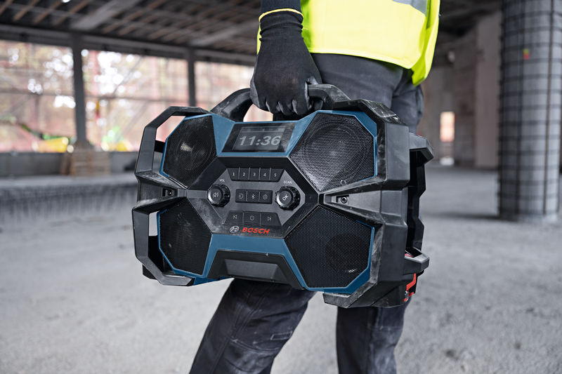 Eine Person mit Sicherheitsausrüstung trägt auf einer Baustelle ein robustes Baustellenradio mit sich. Anwendungsbild