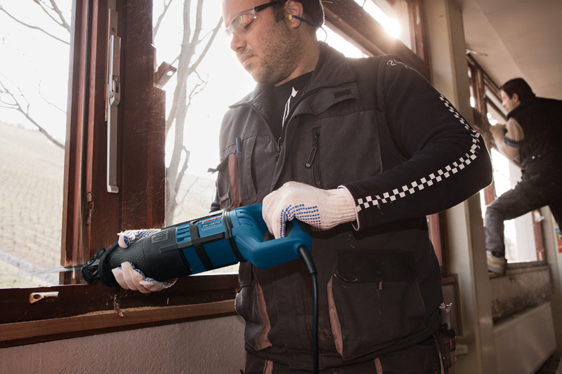 Eine Person mit Sicherheitsausrüstung schneidet mit einer Säbelsäge einen Fensterrahmen durch. Anwendungsbild