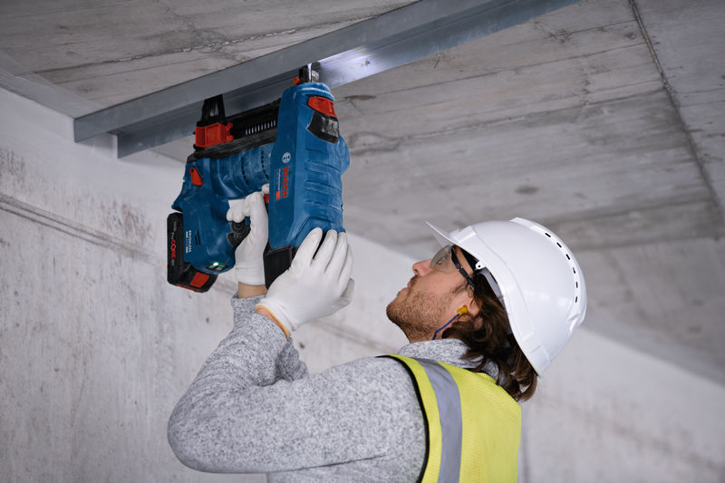 Eine Person mit Sicherheitsausrüstung befestigt mit einem kabellosen Betonnagler einen Metallrahmen an einer Decke. Anwendungsbild