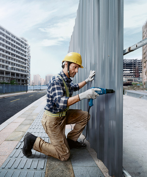 Eine Person mit Sicherheitsausrüstung bohrt auf einer Baustelle in eine Metallwandplatte. Anwendungsbild