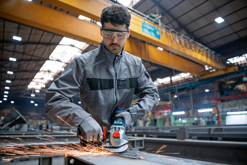 Eine Person mit Schutzausrüstung schleift in einer Fabrik Metall mit einem Akku-Winkelschleifer. Anwendungsbild