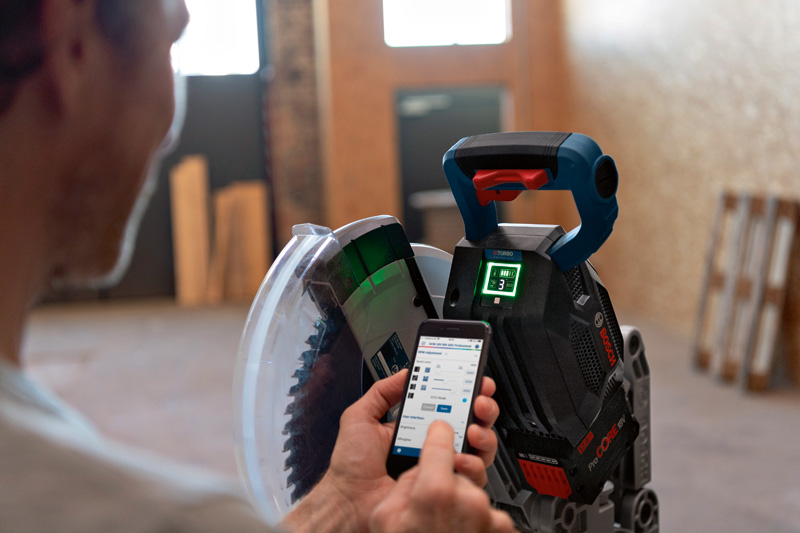 Eine Person stellt in einer Indoor-Werkstatt eine Akku-Gehrungssäge mit einem Smartphone ein. Anwendungsbild