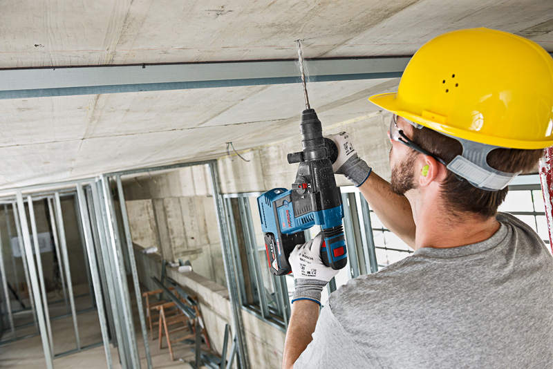Eine Person mit Sicherheitsausrüstung bohrt während der Bauarbeiten in eine Betondecke. Anwendungsbild