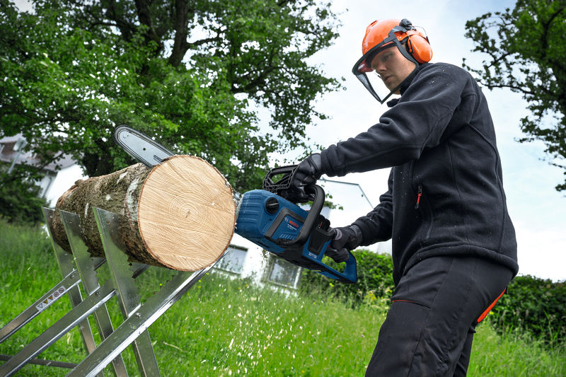 Eine Person mit Sicherheitsausrüstung verwendet eine Kettensäge, um im Freien einen Baumstamm zu zersägen. Anwendungsbild