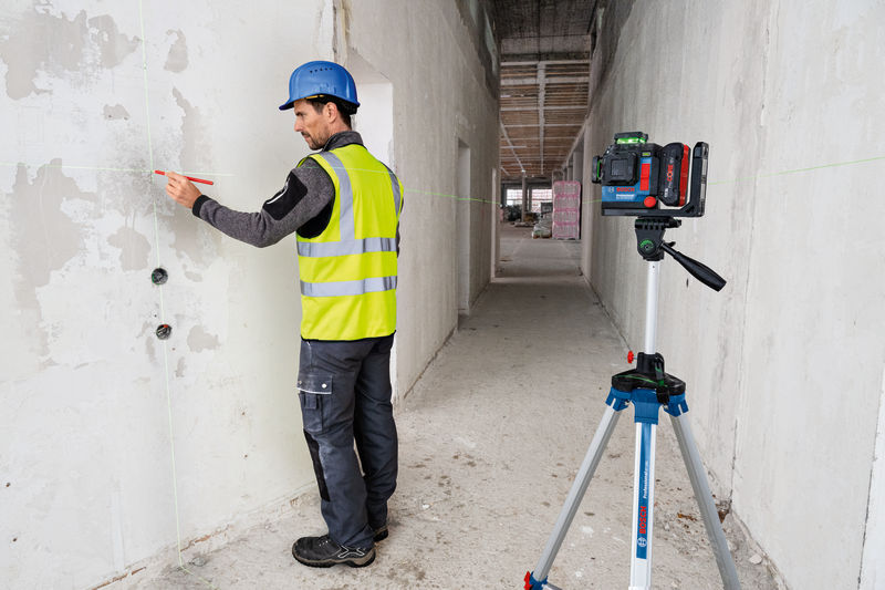 Eine Person mit Sicherheitsausrüstung markiert eine Wand mithilfe eines Lasernivelliergeräts auf einem Stativ. Anwendungsbild