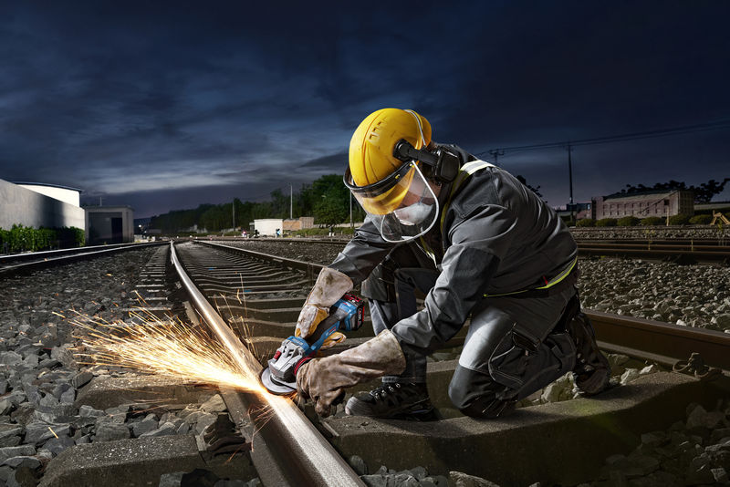 Eine Person mit Sicherheitsausrüstung schneidet mit einem Winkelschleifer ein Eisenbahngleis. Anwendungsbild