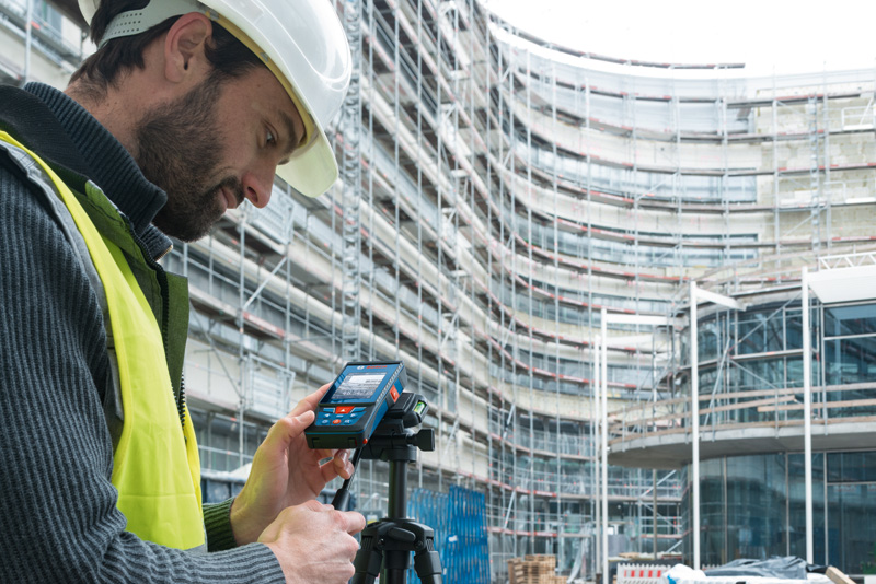 Eine Person mit Sicherheitsausrüstung bedient auf einer Baustelle ein Lasermessgerät. Anwendungsbild