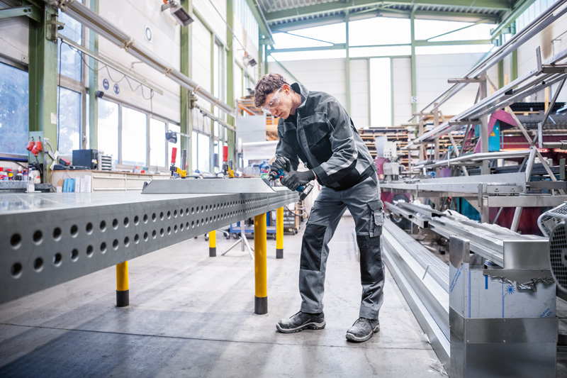 Eine Person mit Schutzausrüstung schleift in einer Werkstatt Metallkanten an einem Lochbalken. Anwendungsbild