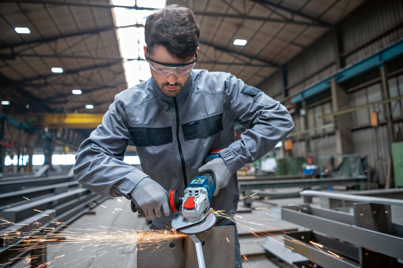 Eine Person mit Schutzausrüstung schleift in einer Fabrik Metall mit einem Akku-Winkelschleifer. Anwendungsbild
