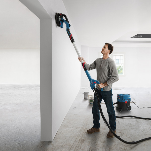 Eine Person schleift eine weiße Wand mit einem Trockenbauschleifer, der an eine Staubabsaugung angeschlossen ist. Anwendungsbild