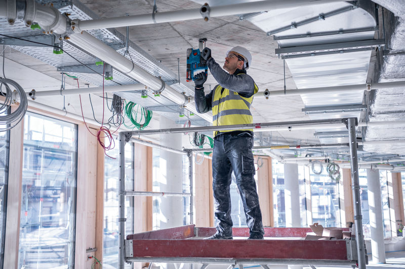Eine Person mit Sicherheitsausrüstung bohrt auf einer Baustelle in eine Betondecke. Anwendungsbild