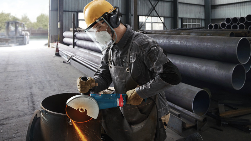 Ein Arbeiter mit Schutzausrüstung schleift mit einem großen Winkelschleifer ein Metallrohr. Anwendungsbild